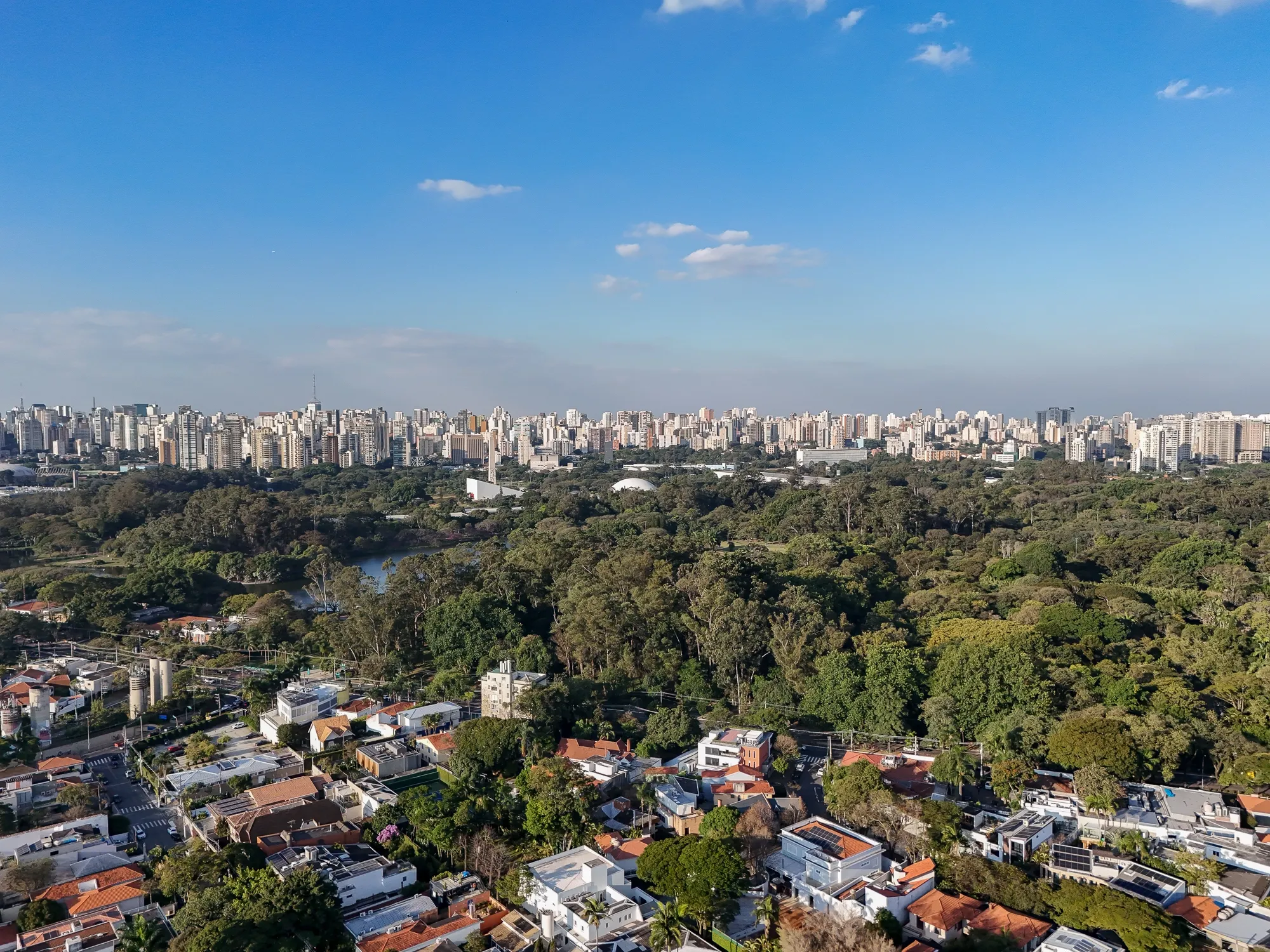 Aerial view of a sprawling city skyline beyond Ibirapuera park, with houses in the foreground under a bright blue sky
