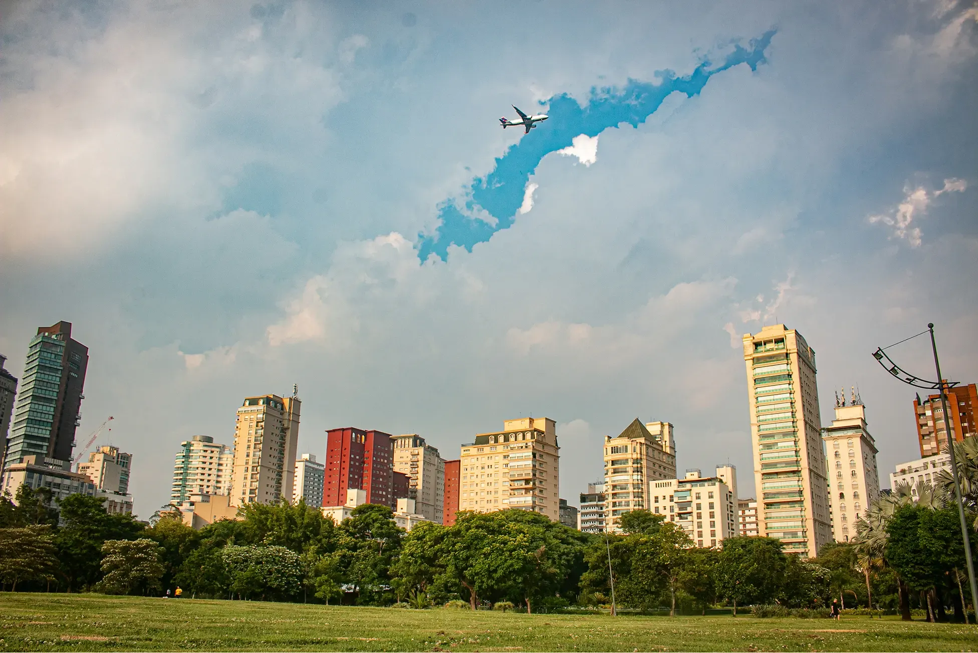 City skyline viewed across a green park under dramatic clouds, with a plane flying through a patch of blue sky
